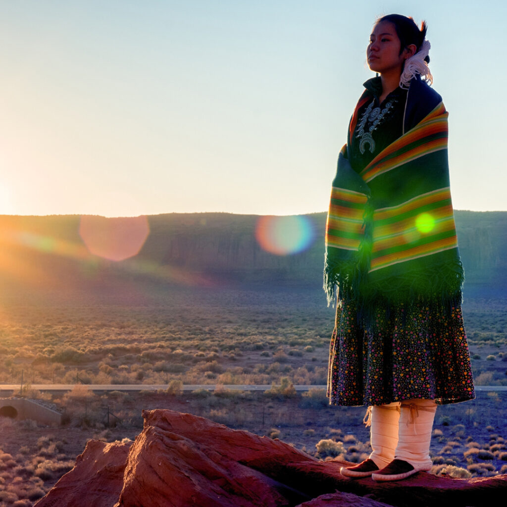Indigenous woman in traditional dress standing on a rock, looking out with a thoughtful expression.