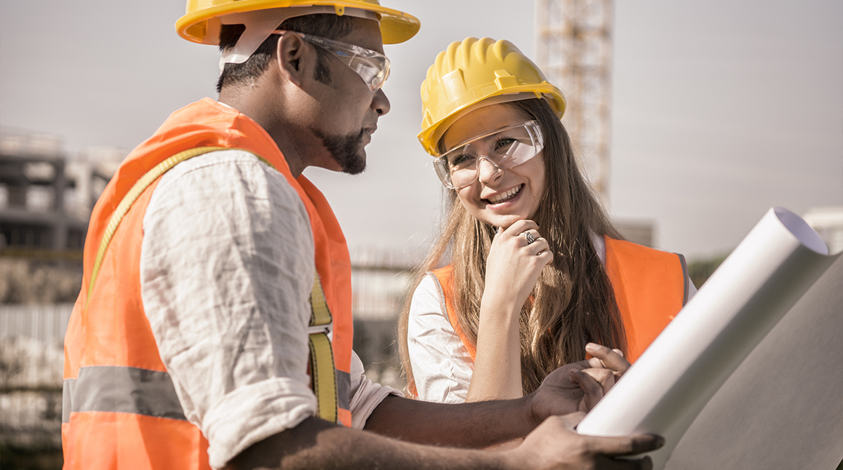 Two workers smiling and chatting, wearing orange safety vests and yellow hardhats.