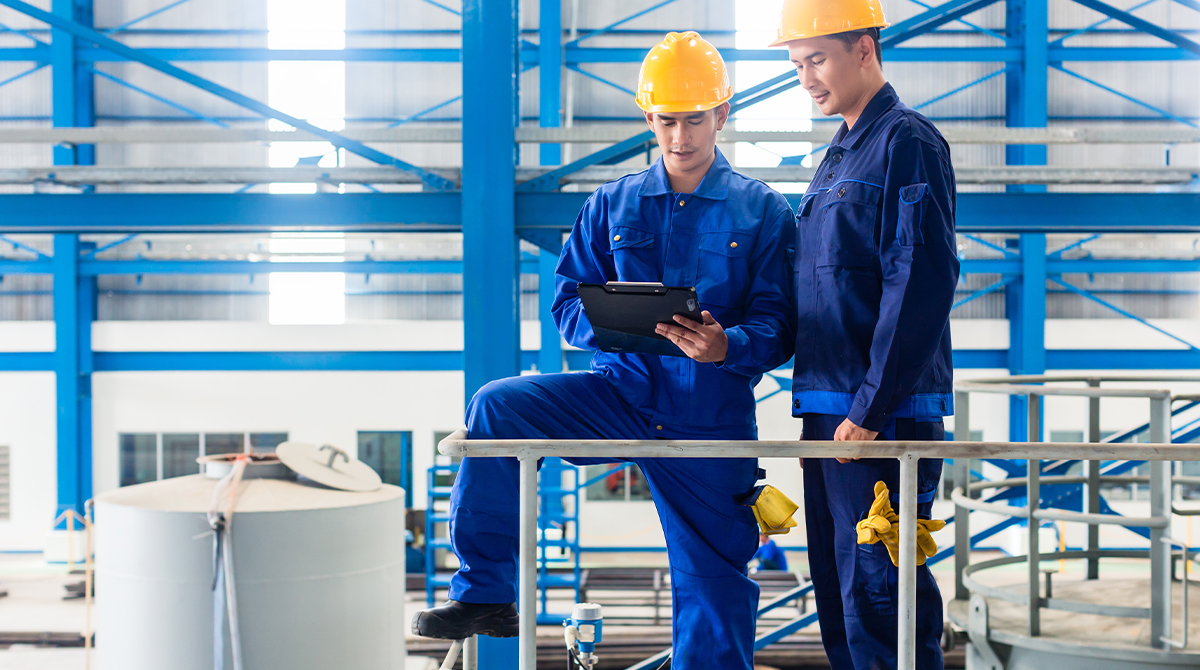 Two workers looking at a clipboard wearing blue coveralls and yellow hardhats.